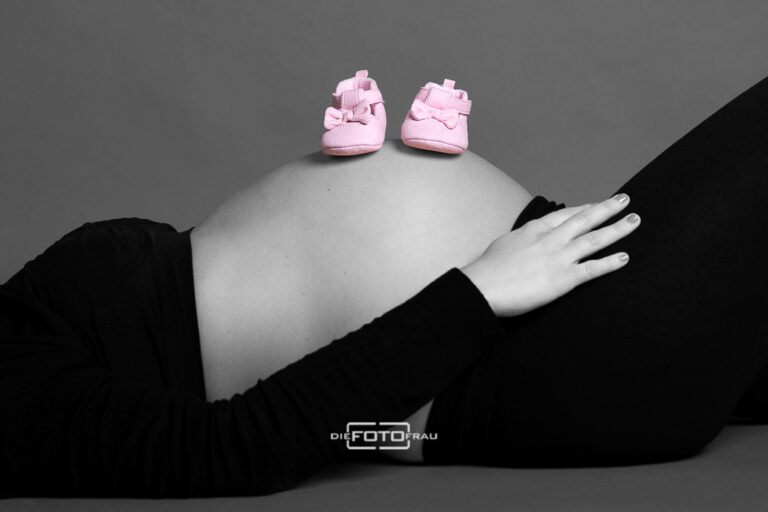 Babybauchshooting im Fotostudio, die werdende Mutter liegt am Boden und auf dem Bauch stehen zwei rosa Babyschuhe