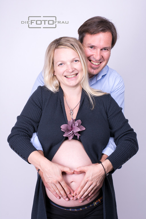Babybauchshooting mit Partner in Alltagskleidung, die Beiden bilden auf dem Bauch ein Herz mit ihren Händen