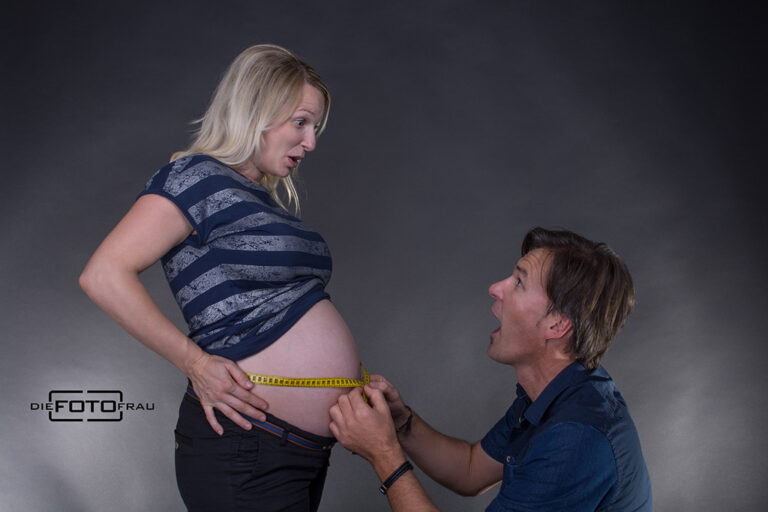 Babybauchshooting als lustiges Foto, wo der Vater den Bauchumfang der werdenden Mutter mit einem Maßband misst
