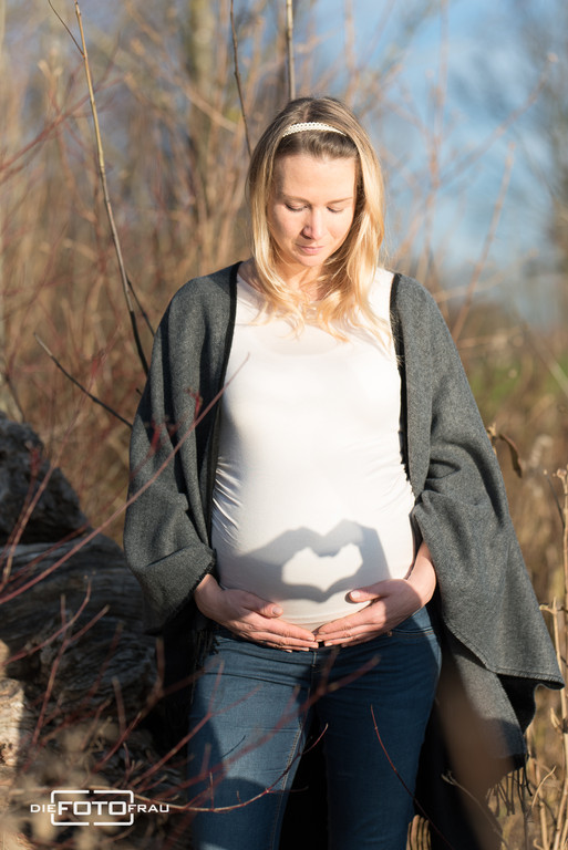 Babybauchshooting im Spätherbst, die werdende Mutter steht im hohen Gras und der Vater zeichnet ein Herz auf den Bauch als Schattenspiel