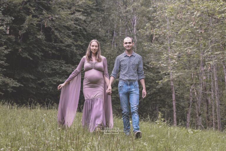 Babybauchshooting mit Partner auf Waldwiese, die werdende Mutter trägt ein violettes Babybauchkleid