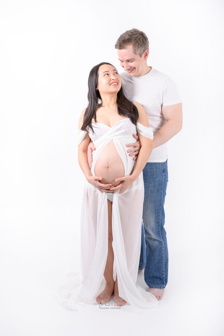 Babybauchshooting mit Partner im Fotostudio, die werdende Mutter trägt ein vorne offenes weißes Dessouskleid