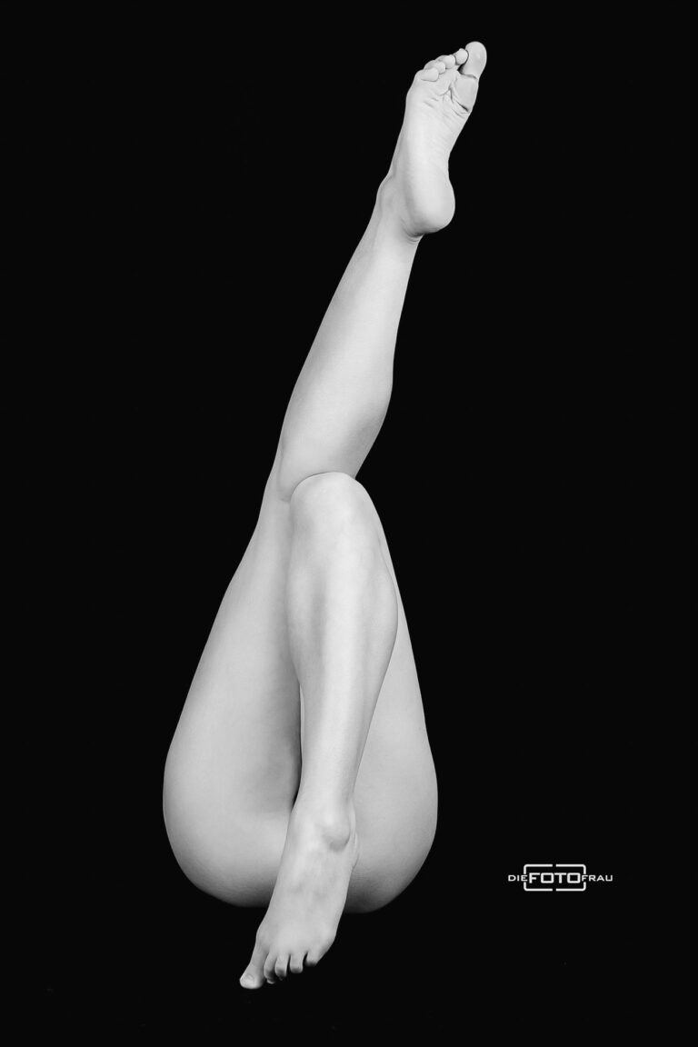 Boudoirfoto in künstlerischer Pose, die Dame streckt einen Fuss in die Höhe und verdeckt mit dem anderen Fuss ihren Po, schwarzweiss