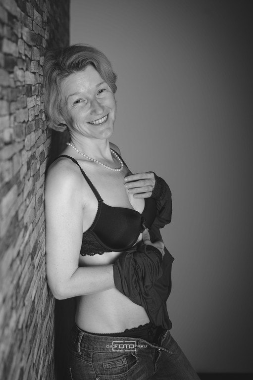 Boudoirfoto in schwarzweiss im Fotostudio Linz mit einer Dame, die an einer Ziegelmauer lehnt und einen schwarzen Büstenhalter trägt