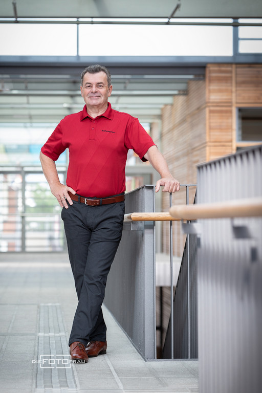 Businessportrait eines Herrn in rotehm Hemd neben einem Metallgelaender in einem Firmengebaeude