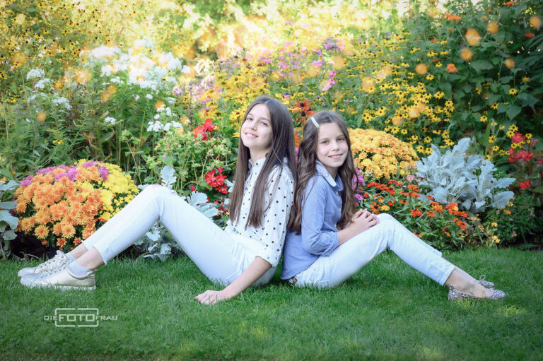 Familienfoto mit Schwestern, diese sitzen auf der Wiese und dahinter sieht man ein Blumenbeet