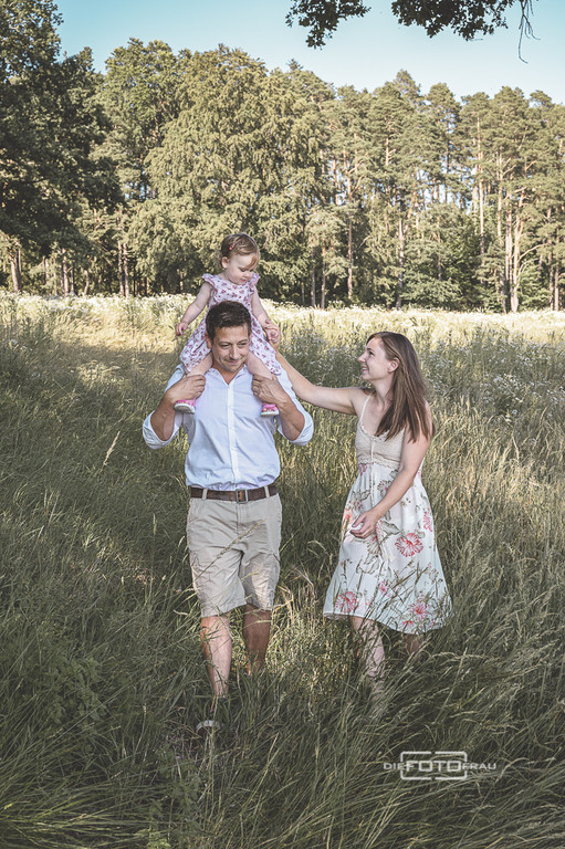 Familienfoto mit Eltern, Vater trägt die kleine Tochter auf den Schultern und alle gehen über eine Gräserwiese