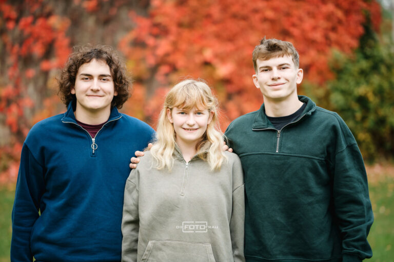 Familienfoto von drei Geschwistern vor einer herbstlich gefärbten Efeumauer