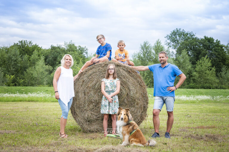 Familienfoto auf einem runden Strohballen, Kinder sitzen darauf und die Eltern stehen links und rechs daneben, ein Collie ist auch dabei