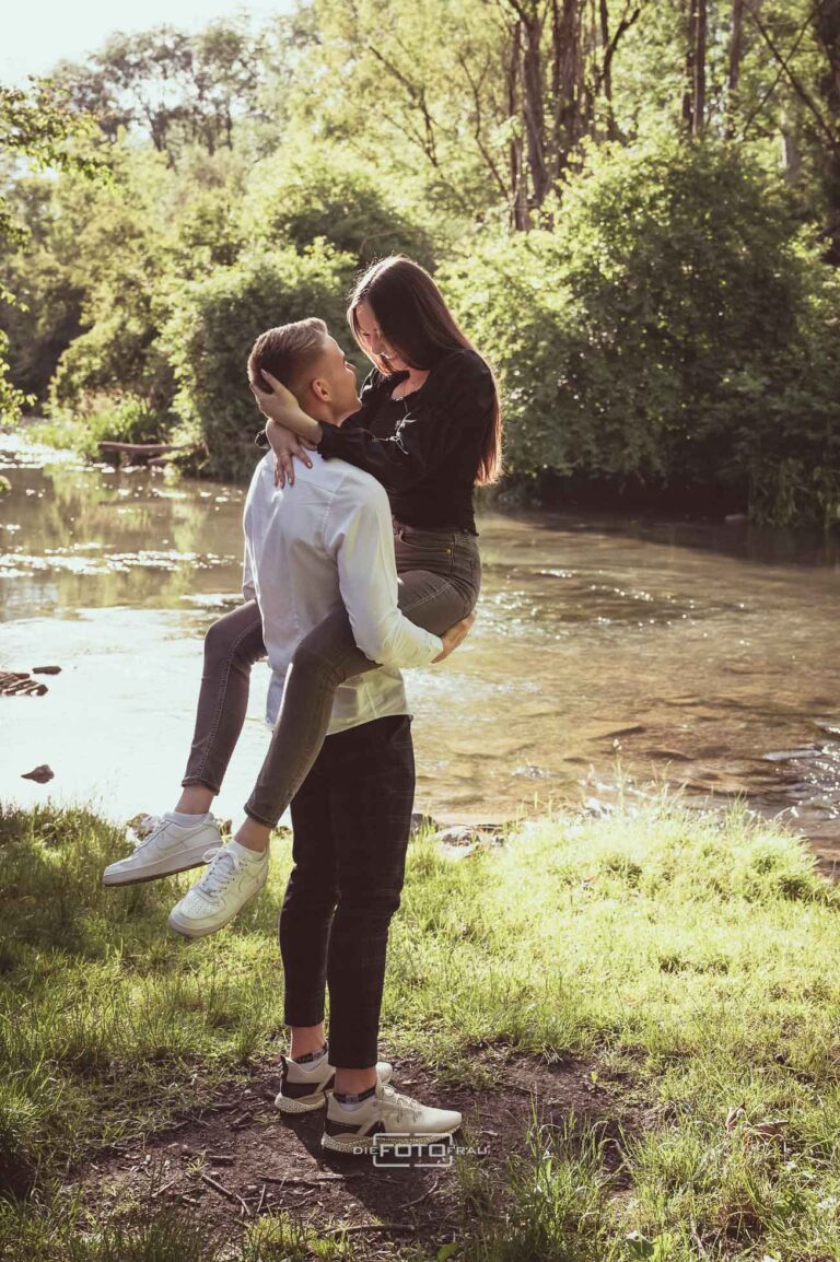 Paarfoto, der Mann trägt die Frau und dahinter sieht man eine Flusslandschaft