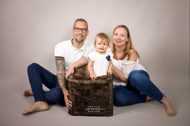 Familienfoto mit Eltern und einjährigem Kind, dieses steht in einer Plüschtruhe