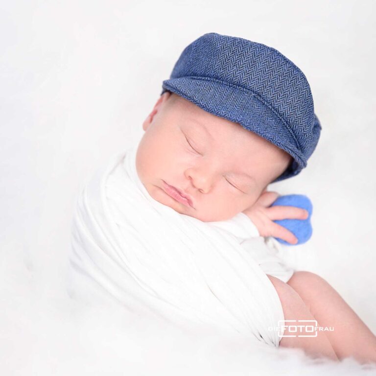 Neugeborenenfoto mit schlafendem Baby, dieses trägt eine blaue Mütze und hält ein blaues Herz in der Hand