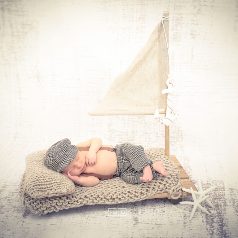 Neugeborenenfoto mit schlafendem Baby auf einem Segelboot, das Baby trägt eine Mütze und eine Latzhose