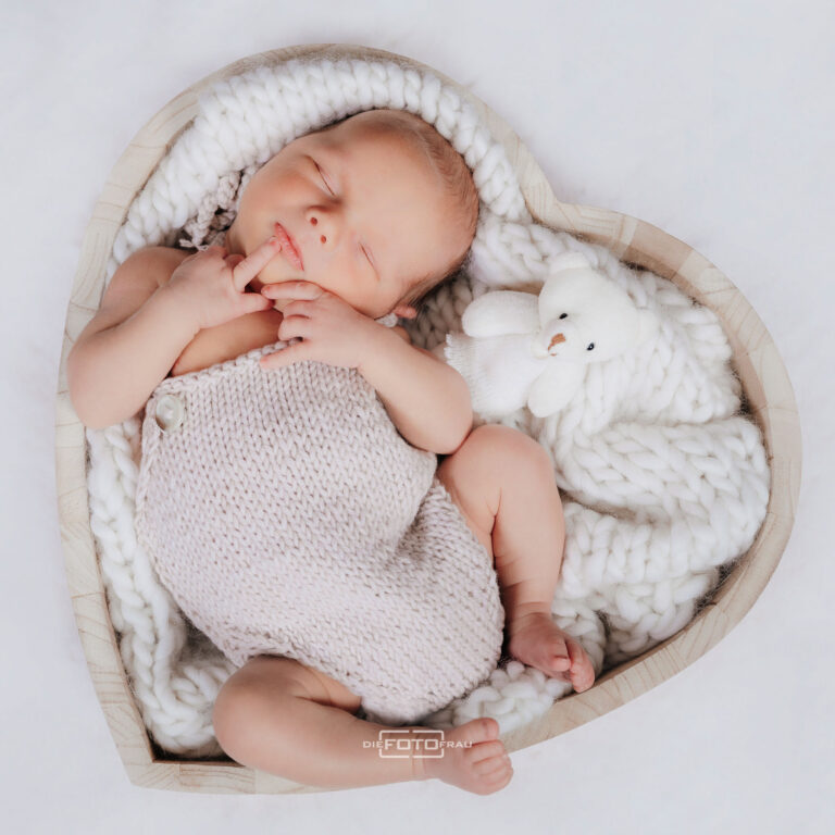 Neugeborenenfoto mit schlafendem Baby in einer herzförmigen Holzschale, das Baby trägt einen handgestrickten Body