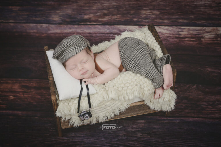 Neugeborenenfoto mit schlafendem Baby in einem Holzbett, dieses hält eine Spielzeugkamera in der Hand und trägt Mütze und Latzhose