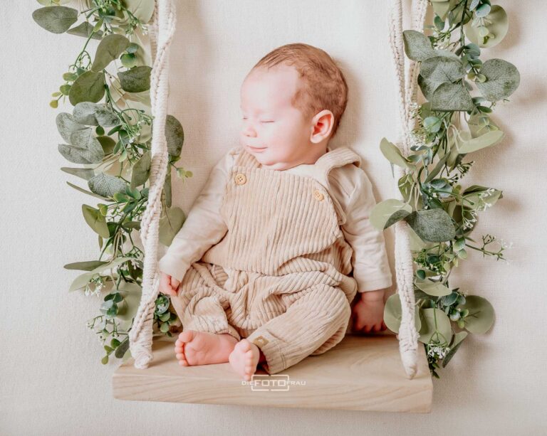 Neugeborenenfoto mit Baby in Latzhose auf Schaukel mit Eukalyptus dekoriert