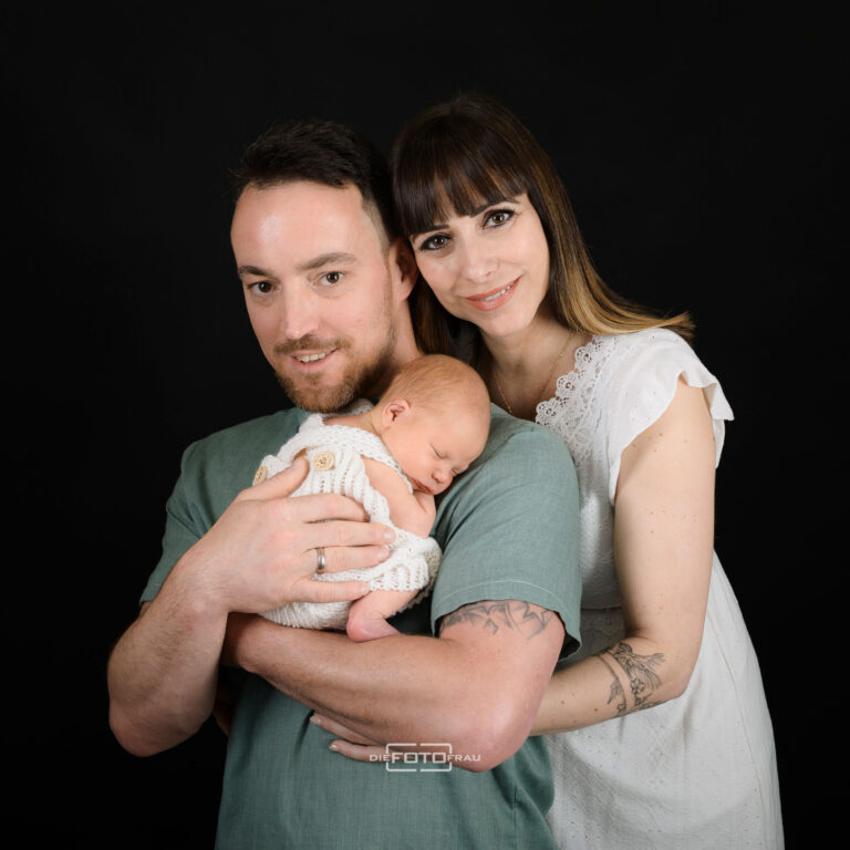 Neugeborenenfoto mit schlafendem Baby in Papas Armen, die Mutter umarmt die Beiden