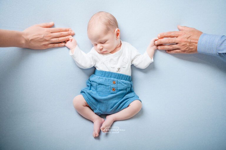 Neugeborenenfoto mit schlafendem Baby auf blauem Untergrund, die Eltern halten jeweils eine Babyhand