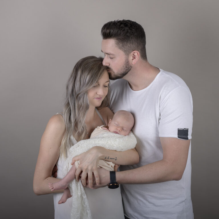 Neugeborenenfoto mit schlafendem Baby und den Eltern, der Papa küsst die Mama