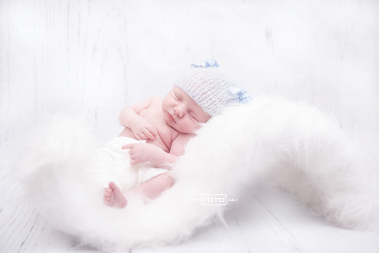 Neugeborenenfoto mit schlafendem Baby auf Fellwiege, weisser Hintergrund und blaue Babymütze
