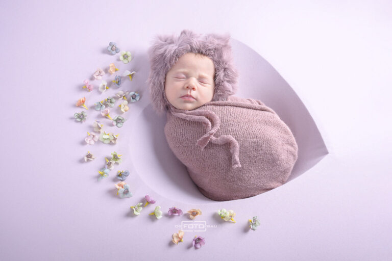 Neugeborenenfoto mit schlafendem Baby in einer Herzschale, dekoriert mit Blüten und rosa Mütze und Tüchern