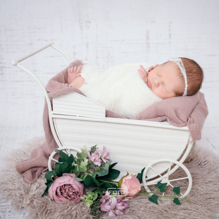 Neugeborenenfoto mit schlafendem Baby in einem weißen Kinderwagen und Blumendeko