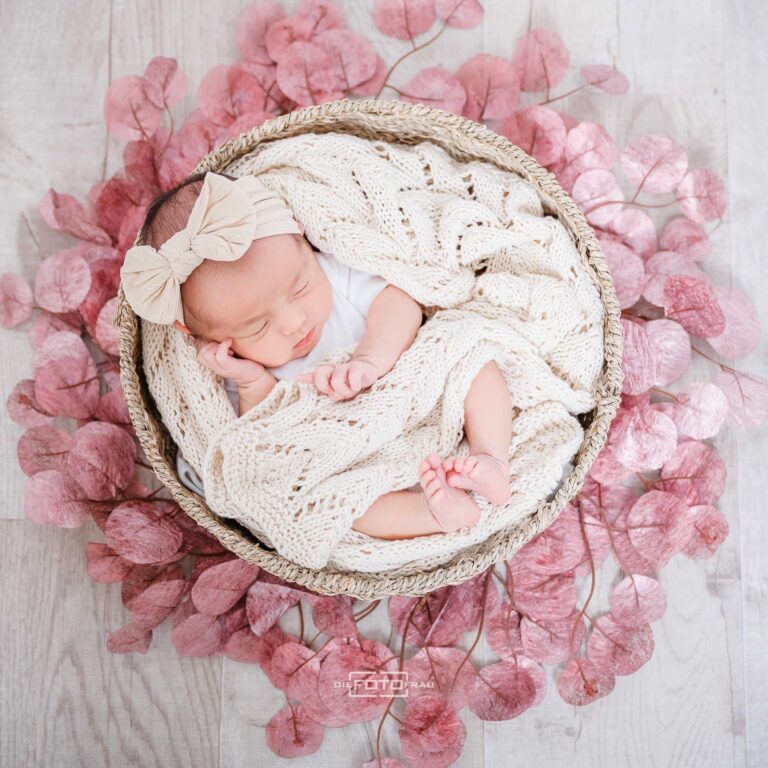 Neugeborenenfoto, das Baby liegt auf Tücher gebettet in einem runden Strohkorb mit rosa Blütenblättern