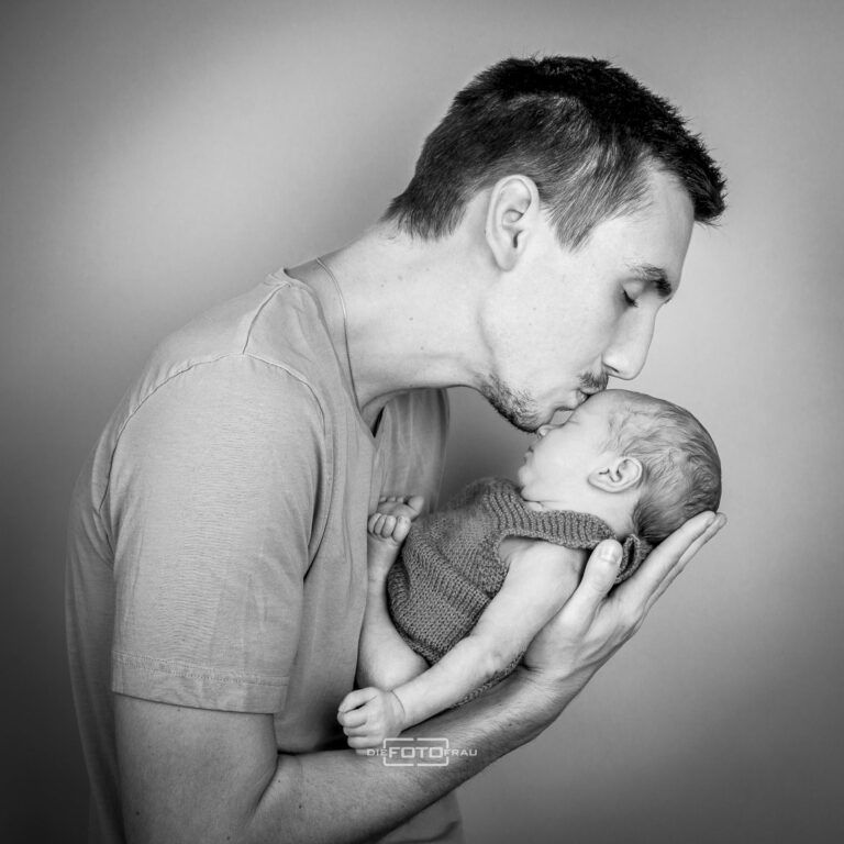 Neugeborenenfoto mit schlafendem Baby in Papas Armen, dieser küsst das Baby