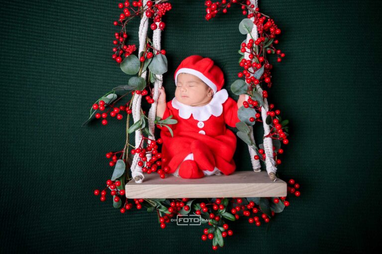 Neugeborenenfoto mit schlafendem Baby in weihnachtlich dekorierter Schaukel, das Baby trägt ein rotes Weihnachtskleid mit Mütze