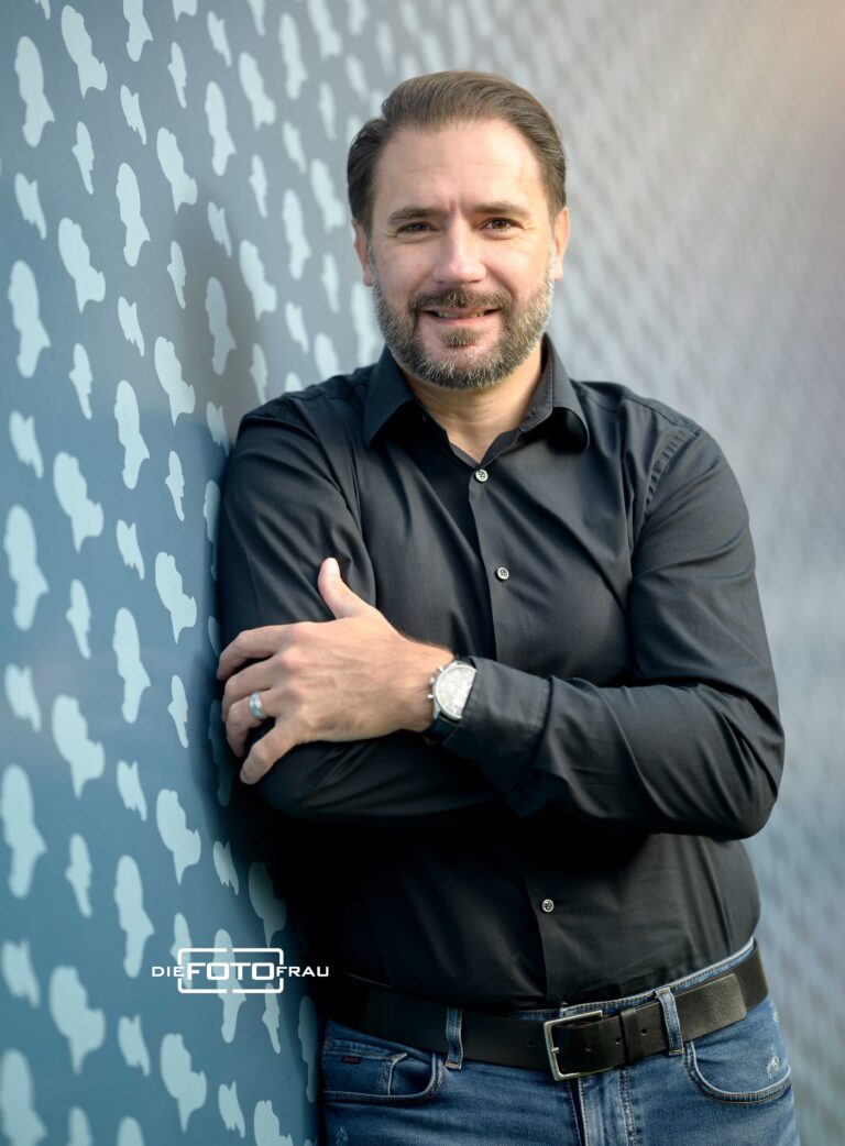 Portraitfoto eines dunkelhaarigen Mannes im schwarzen Hemd vor einer bunten Wand