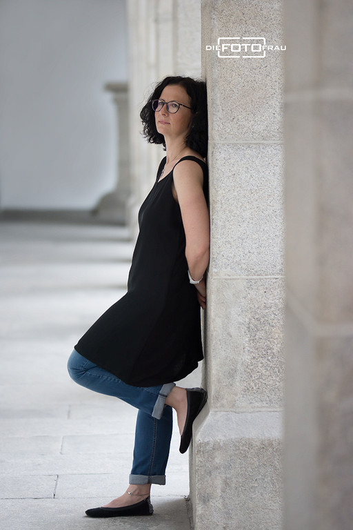 Portraitfoto einer Dame an eine Granitsäule gelehnt, sie trägt ein schwarzes Kleid und Jeans