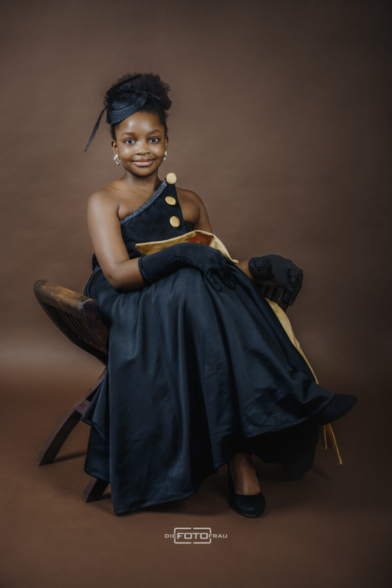 Portraitfoto eines jungen Mädchens aus Ghana im Festtagskleid mit Kopfschmuck und Handschuhen, auf einem Stuhl im Fotostudio Linz sitzend