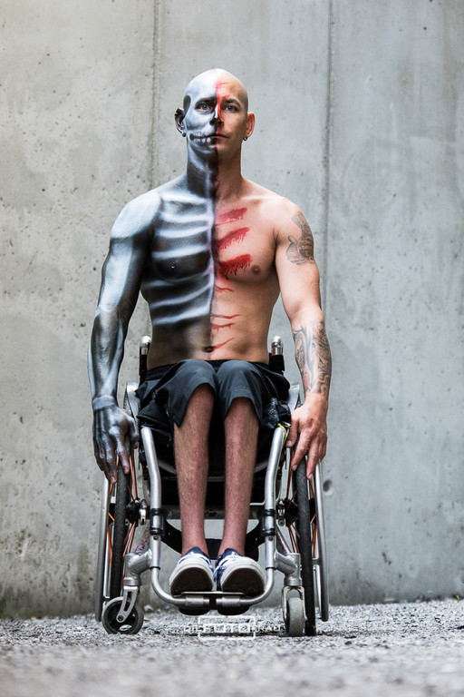 Portraitfoto eines Herrn im Rollstuhl sitzend vor einer Betonmauer, er trägt eine Bodypainting Bemalung