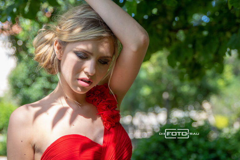 Portraitfoto einer jungen Dame mit rotem Kleid unter einem Baum stehend, die Blätter werfen dekorative Schatten auf das Dekollete