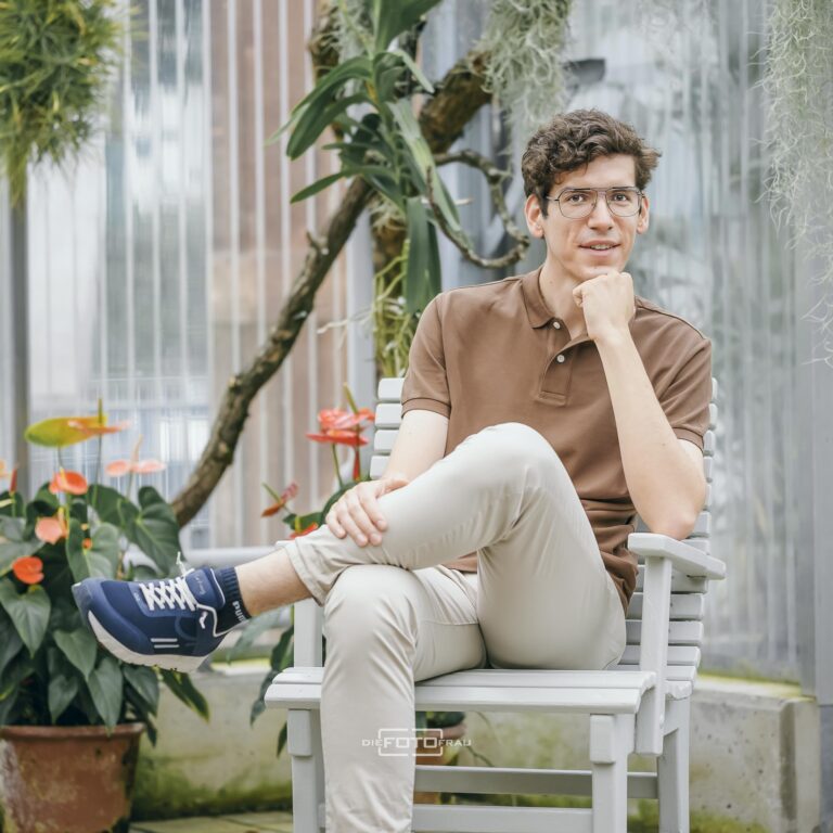 Portraitfoto eines jungen Mediziners in Freizeitkleidung im Botanischen Garten in Linz, auf einem Stuhl sitzend