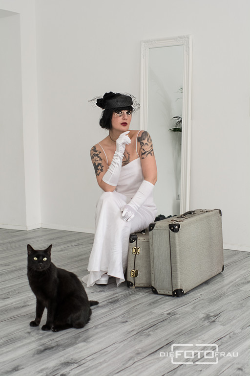 Portraitfoto einer Dame im Reiseoutfit mit Koffer, davor sitzt eine schwarze Katze