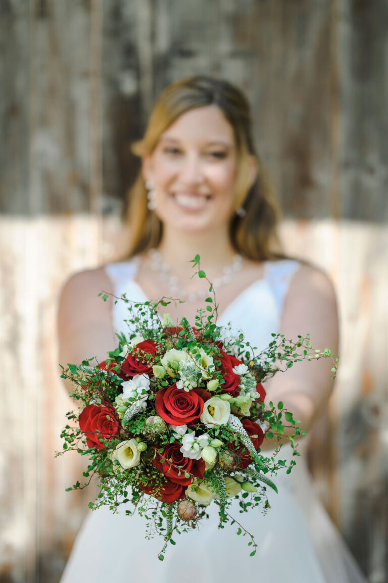 Braut hält stolz ihren Brautstrauss mit roten Rosen und weißen Blüten vor sich