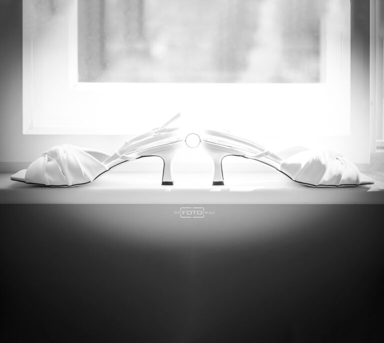 Weiße Hochzeitsschuhe stehen mit den Fersen gegeneinander auf einem Fensterbrett, zwischen den Fersen befindet sich der Ehering,schwarzweisses Foto