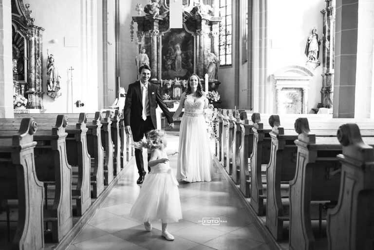 Brautpaar beim Auszug aus der Kirche, die Tochter läuft vor ihnen und blickt auf die Eltern zurück, schwarzweisses Foto