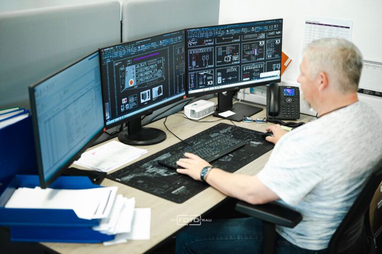Mitarbeiter am Schreibtisch vor 3 Computern mit technischen Zeichnungen