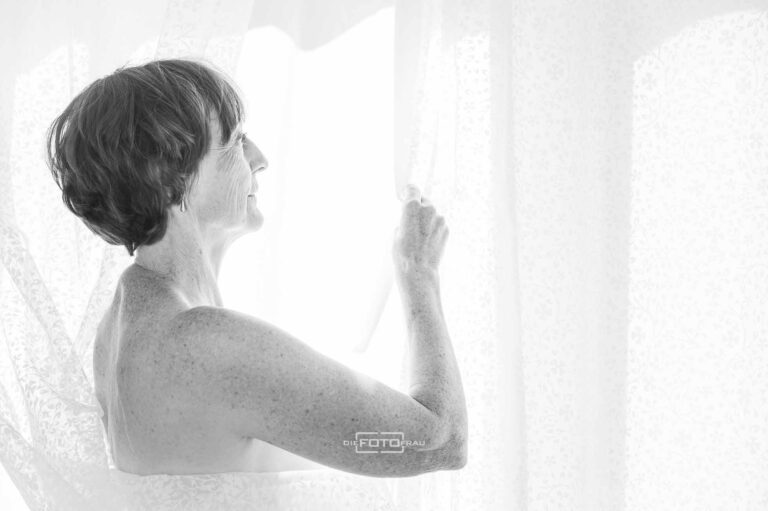 Reifere Dame in weisses Tuch gehüllt blickt durch Vorhang aus dem Fenster, schwarzweisse Aufnahme