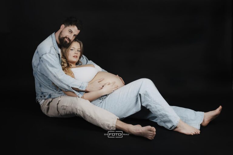 Schwangere Frau liegt in den Armen ihres Partners, beide tragen Jeanslook, aufgenommen im Fotostudio DieFotoFrau